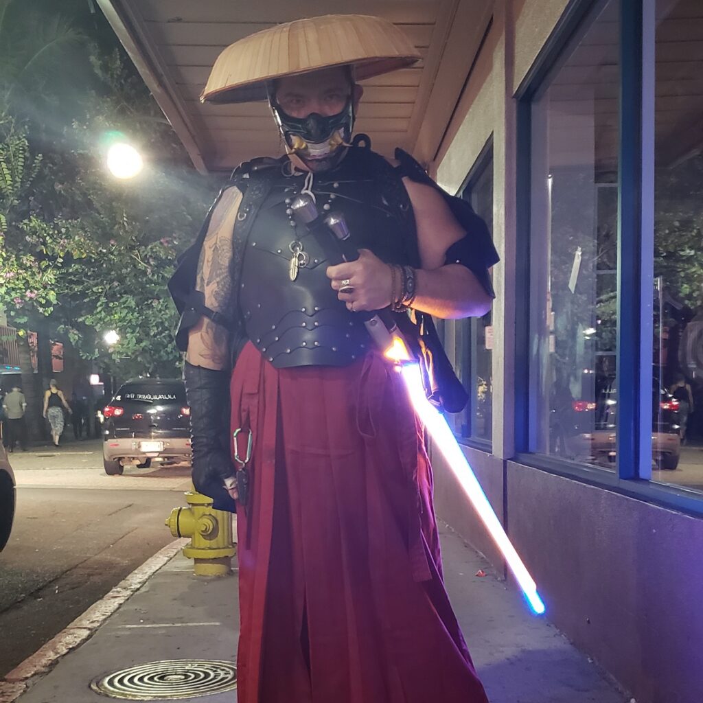 Person in samurai-style armor with a conical hat and mask, holding a glowing blue lightsaber on a city sidewalk at night.