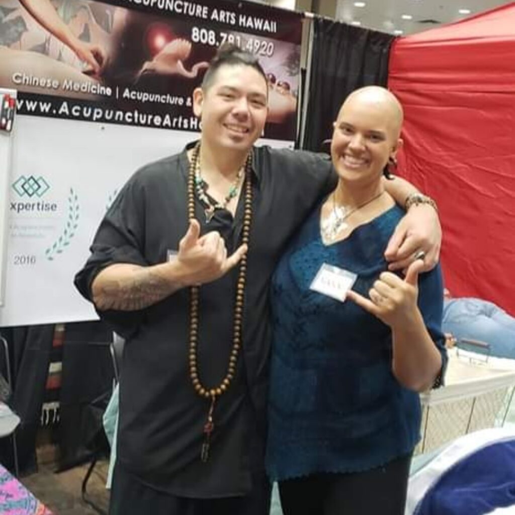 Two people posing together at a booth at a fair; man in black with a beaded necklace making a rock-on gesture, woman in blue top with a name badge, both smiling. Booth banner in background reads Acupuncture Arts Hawaii.