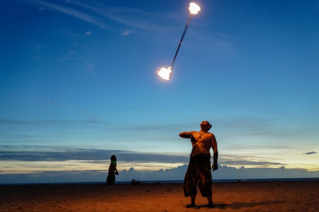 Two fire performers on a beach at dusk, one spinning a long arc of flames across the sky.